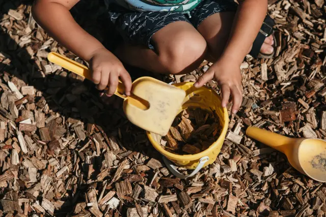 Kinderhände füllen Holzschnitzel mit einer gelben Schaufel in einen Kessel